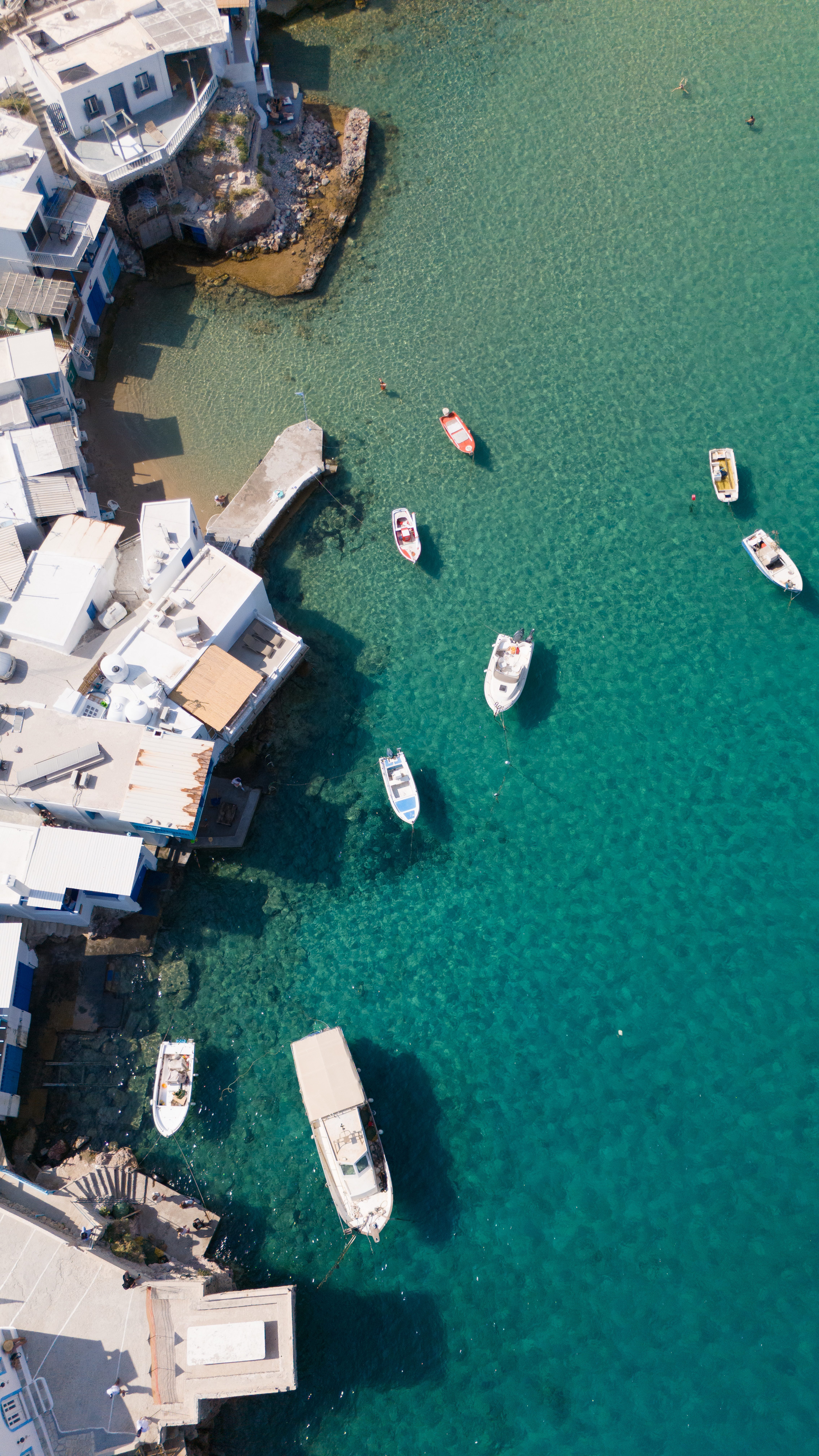 Aerial view of fishing boats in a turquoise harbor beside whitewashed buildings in Greece