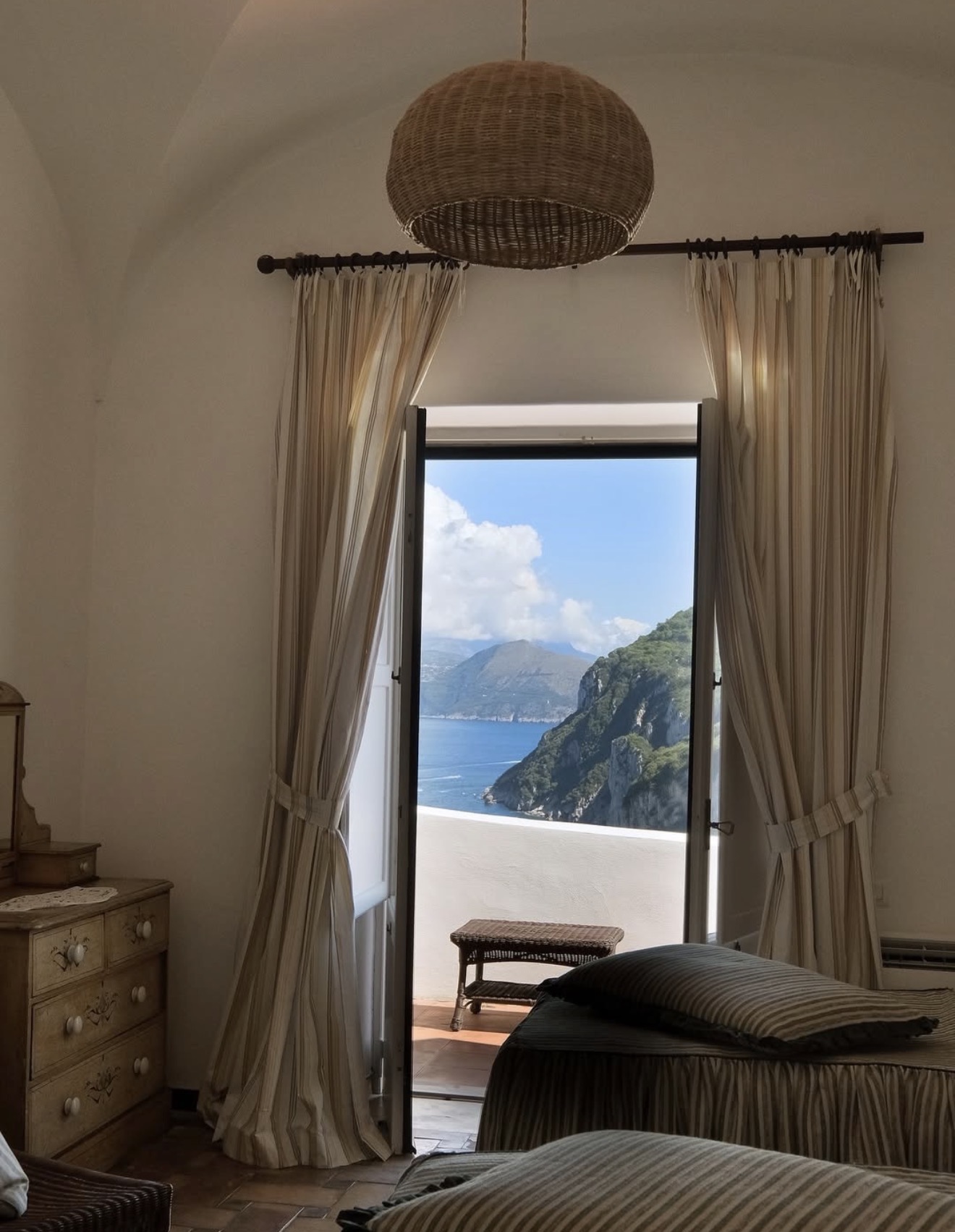 A curtained balcony doorway framing a view of the cliffs and sea along the coast of Capri