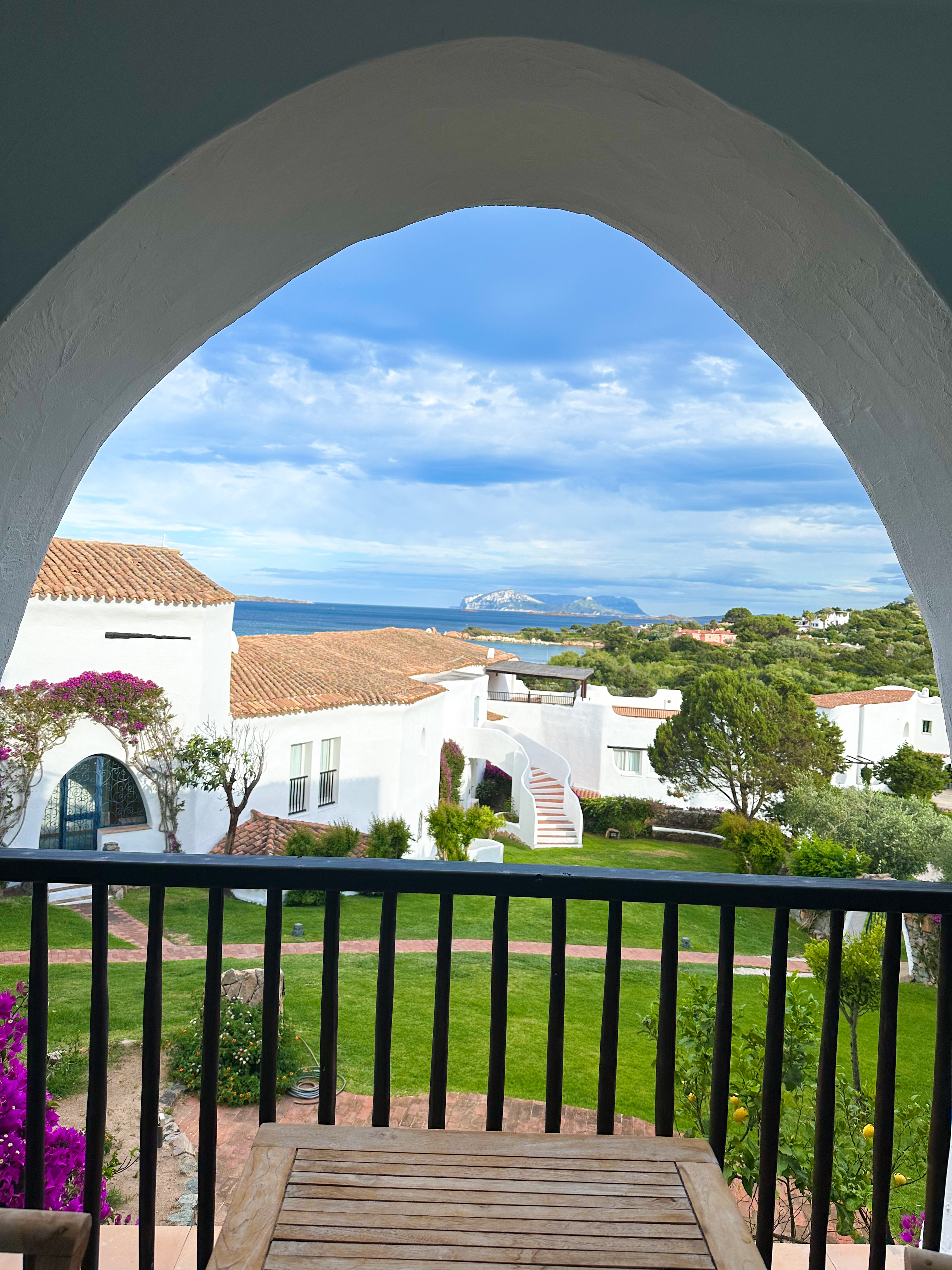 A view through a white archway over villa rooftops, gardens, and the sea along the coast of Sardinia, Italy