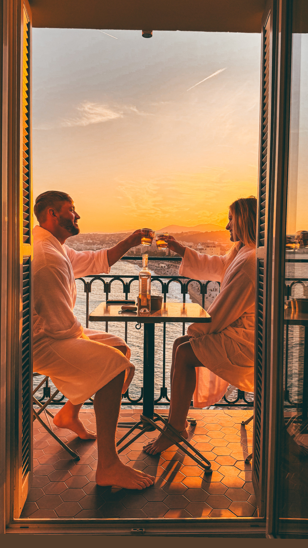 A couple in robes toasting at sunset on a private balcony overlooking the water